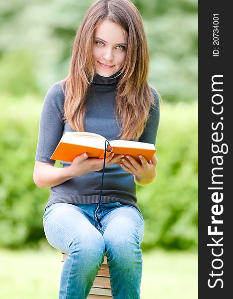 Beautiful and happy young student girl sitting on pile of books, holding book in her hands, smiling and looking into the camera. Summer or spring green park in background. Beautiful and happy young student girl sitting on pile of books, holding book in her hands, smiling and looking into the camera. Summer or spring green park in background