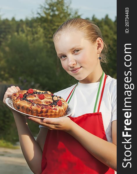 The girl the teenager in a red apron holds in a hand a pie from fresh berries against summer wood. The girl the teenager in a red apron holds in a hand a pie from fresh berries against summer wood