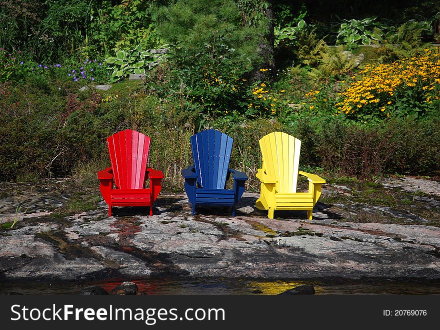 This is a picture of three wooden Beach chairs that are sitting on a rock shoreline. They are three different colors and are in front of a wooded background. This is a picture of three wooden Beach chairs that are sitting on a rock shoreline. They are three different colors and are in front of a wooded background.