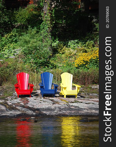 This is a picture of three wooden beach chairs all of different colors. They have been situated on a beautiful rock shoreline of a lake with a wooded background. One red, one blue, and one yellow chair. This is a picture of three wooden beach chairs all of different colors. They have been situated on a beautiful rock shoreline of a lake with a wooded background. One red, one blue, and one yellow chair.