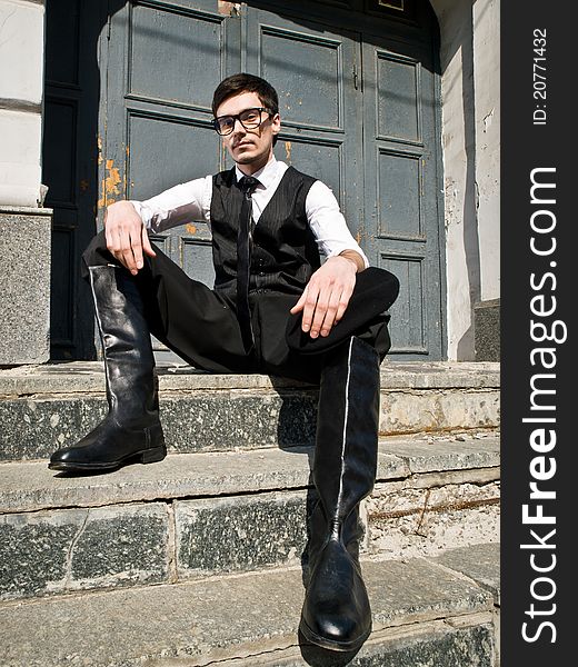 Young man in eyeglass and boots in vintage style is sitting against the background old door. Young man in eyeglass and boots in vintage style is sitting against the background old door