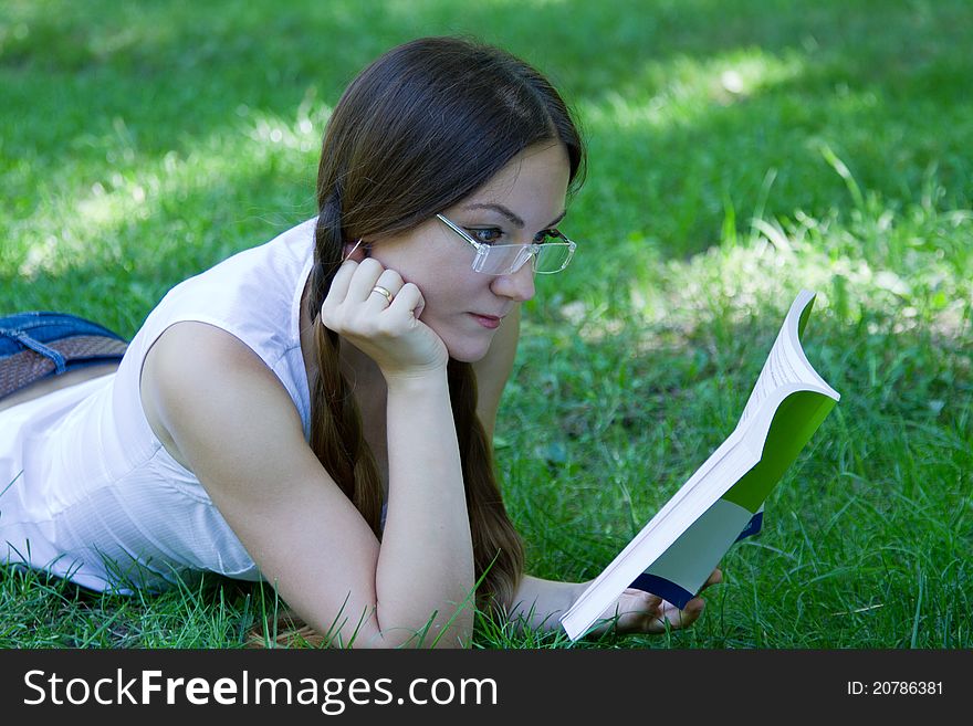 Beautiful student with two braids wearing jeans lying on the grass with a book in the park. Beautiful student with two braids wearing jeans lying on the grass with a book in the park