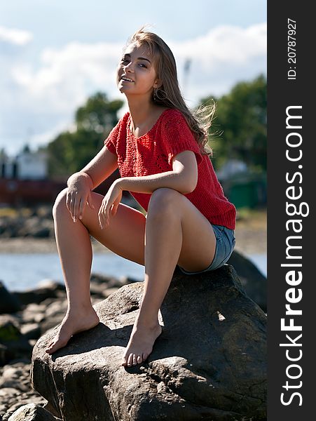 Girl in a red sweater sits on a rock against the backdrop of the coastline