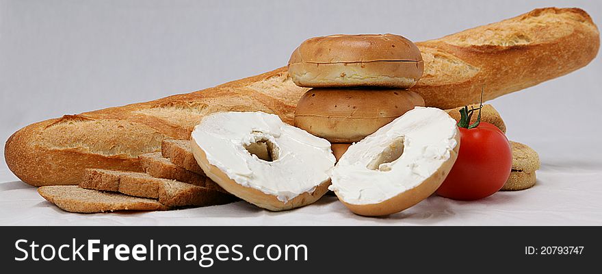 Bagel opened and spread with cream cheese in front of a variety of bread and a tomato. Bagel opened and spread with cream cheese in front of a variety of bread and a tomato.