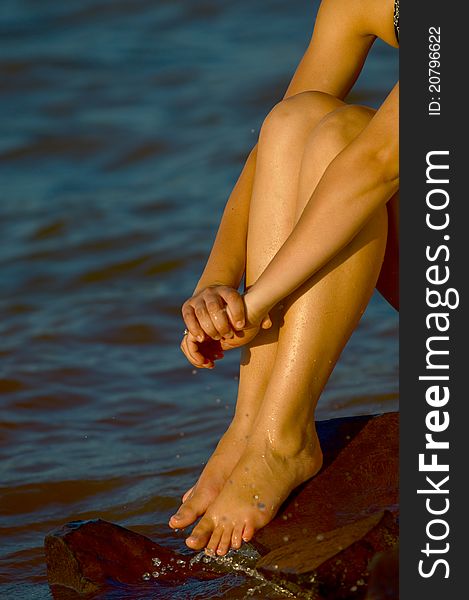 Teen woman in swimsuit sitting on rocks by water at sunset. Teen woman in swimsuit sitting on rocks by water at sunset