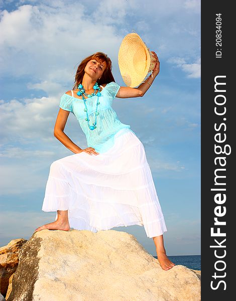 Attractive woman holding his hat (squash) in a hand step on a rock with cloudy sky and blue sea (ocean) background. Attractive woman holding his hat (squash) in a hand step on a rock with cloudy sky and blue sea (ocean) background