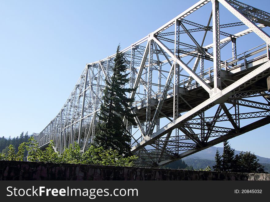 A closeup of an image of the historic Bridge of the Gods in Cascade Locks, Oregon over the Columbia River Gorge with trees in the foreground & blue sky. Selective focus. A closeup of an image of the historic Bridge of the Gods in Cascade Locks, Oregon over the Columbia River Gorge with trees in the foreground & blue sky. Selective focus.