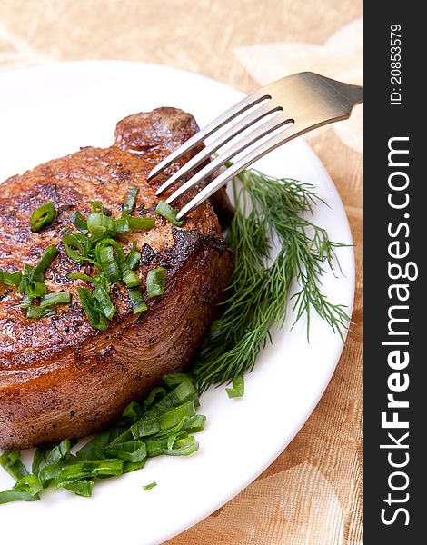 Big juicy well-done piece of meat lying on a white plate decorated with greenery with a fork. Big juicy well-done piece of meat lying on a white plate decorated with greenery with a fork