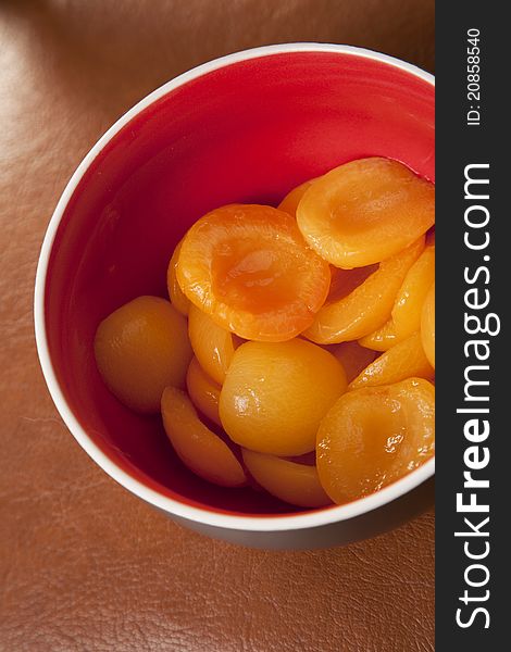 Delicious peaches in syrup sauce in a red bowl, on leather background.