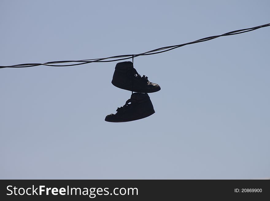 A pair of discarded black shoes hanging from a telephone wire with a blue sky in the background. A pair of discarded black shoes hanging from a telephone wire with a blue sky in the background.