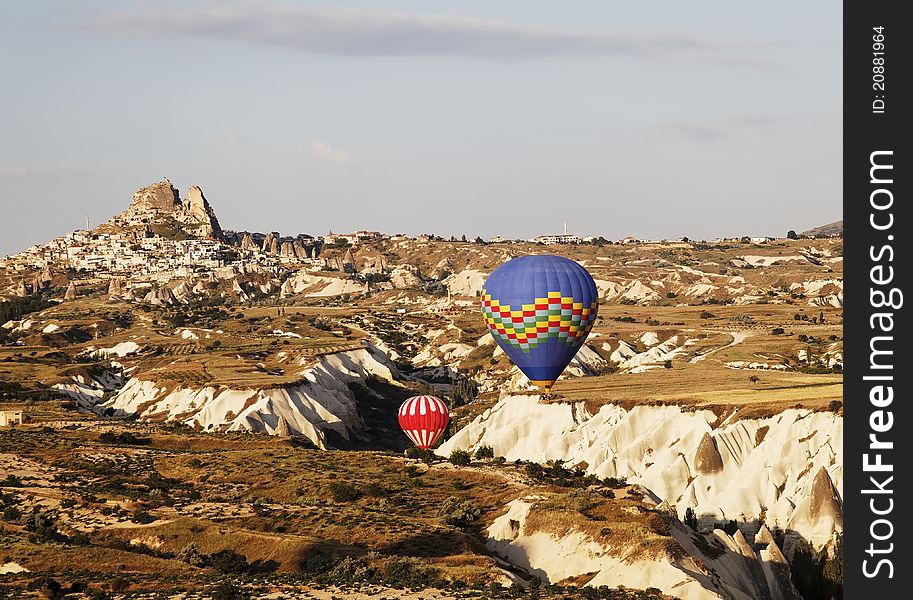 Uchisar backdrop red and white plus blue multi colored hot air balloons navigate a gorge during low level flight, blue sky, wilderness, desert plants, limestone cleft, volcanic region, landscape, crop space and copy area. Uchisar backdrop red and white plus blue multi colored hot air balloons navigate a gorge during low level flight, blue sky, wilderness, desert plants, limestone cleft, volcanic region, landscape, crop space and copy area