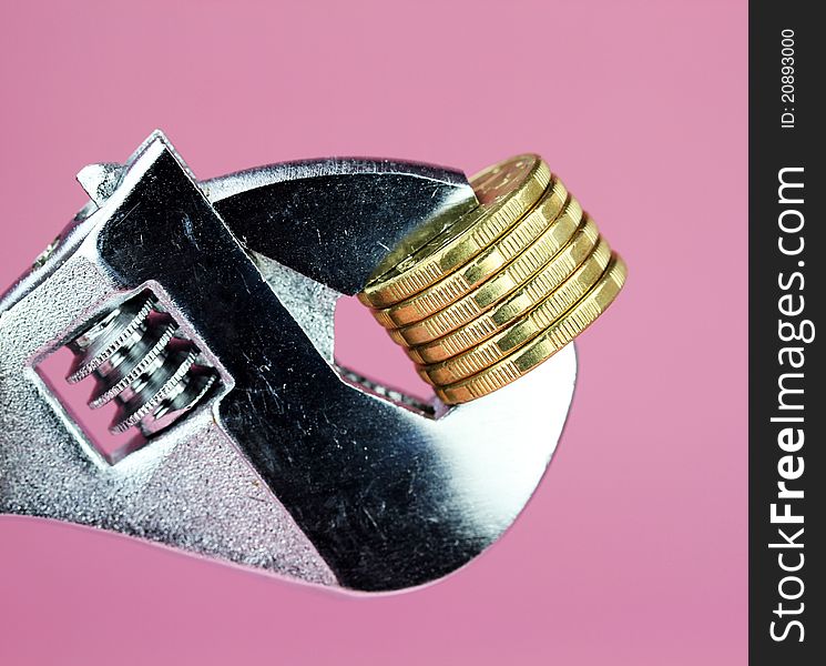 A stack of Gold Dollar Coins being squeezed in a silver adjustable spanner, against a light pastel pink background and asking the questions how tight is you household budget?. A stack of Gold Dollar Coins being squeezed in a silver adjustable spanner, against a light pastel pink background and asking the questions how tight is you household budget?