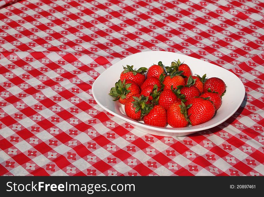 A dish of freshly picked strawberries on a red and white checkered picnic blanket in the sun. A dish of freshly picked strawberries on a red and white checkered picnic blanket in the sun.