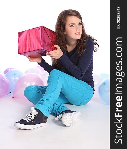 Beautiful teenager girl with bright blue eyes celebrates happy occasion with a surprise birthday present wrapped in pink gift paper, sitting among party balloons. Beautiful teenager girl with bright blue eyes celebrates happy occasion with a surprise birthday present wrapped in pink gift paper, sitting among party balloons.