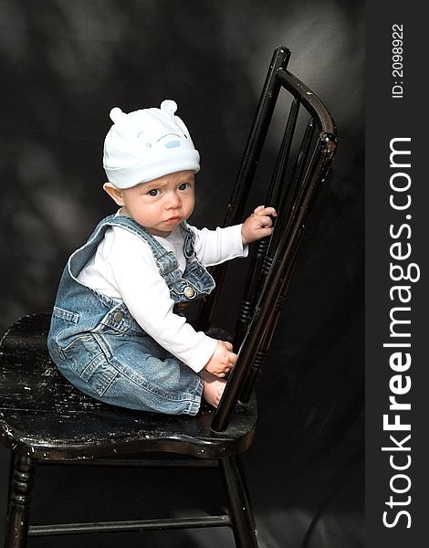 Image of adorable baby wearing denim overalls sitting on a black chair. Image of adorable baby wearing denim overalls sitting on a black chair