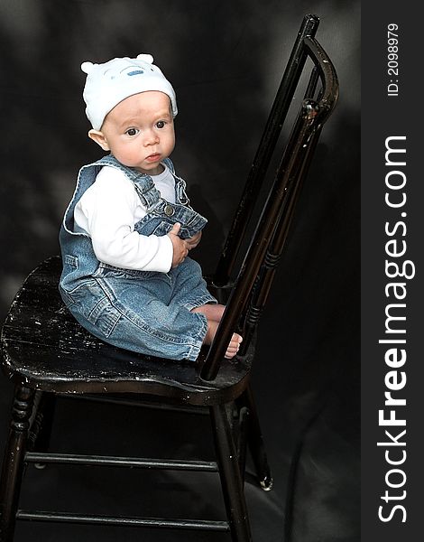 Image of adorable baby wearing denim overalls and a blue cap sitting on a black chair. Image of adorable baby wearing denim overalls and a blue cap sitting on a black chair