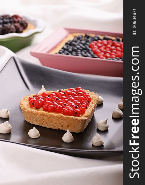 Heart-shaped tart with redcurrants served on a black plate decorated with samll meringues. Tarts with berries out of focus in the background. Selective focus, shallow DOF. Heart-shaped tart with redcurrants served on a black plate decorated with samll meringues. Tarts with berries out of focus in the background. Selective focus, shallow DOF