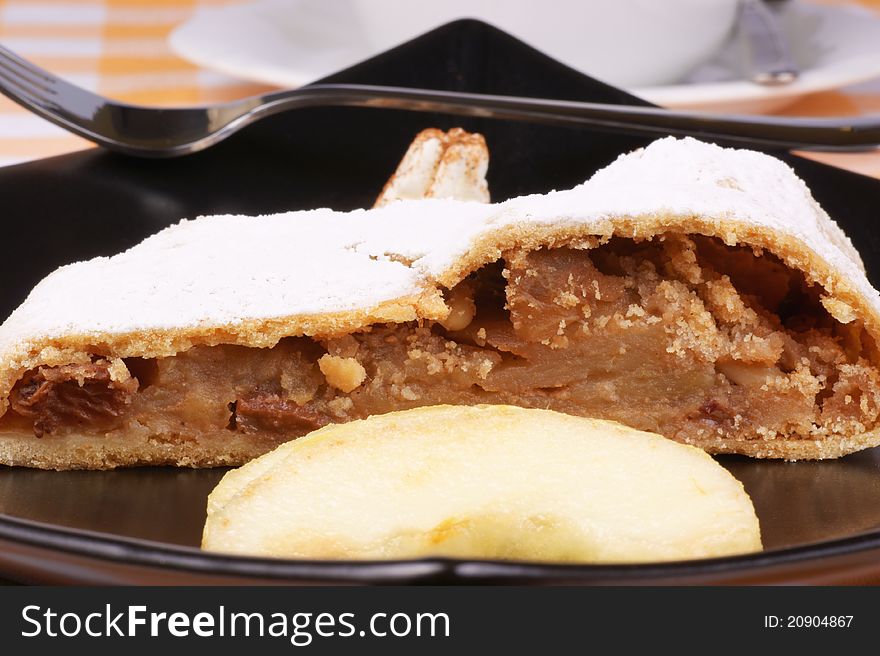 Apple strudel (Apfelstrudel) served on a black plate with a tea cup in the background. Selective focus, shallow DOF. Apple strudel (Apfelstrudel) served on a black plate with a tea cup in the background. Selective focus, shallow DOF.