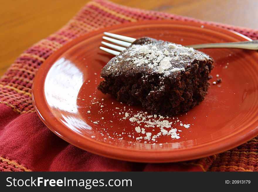 A brownie with powdered sugar on top on a reddish orange plate. A brownie with powdered sugar on top on a reddish orange plate.