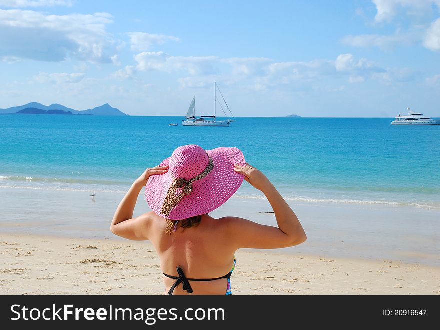 Young woman standing on a beach holding her straw hat and looking to the horizon.