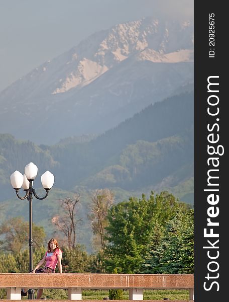 Image of National Park in Almaty. Kazakhstan. Young woman is sitting opposite street lamp with beautiful view on mountain. Image of National Park in Almaty. Kazakhstan. Young woman is sitting opposite street lamp with beautiful view on mountain.
