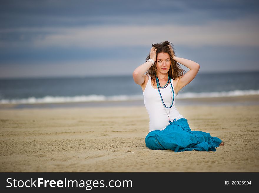 Beautiful woman in white cami and turquoise skirt sits on sand near the ocean. Beautiful woman in white cami and turquoise skirt sits on sand near the ocean