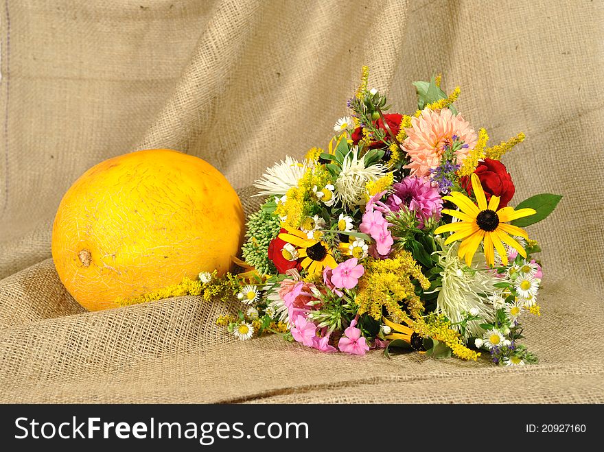 The autumn topic. In the foreground is bouquet of wild and cultivated flowers and orange melon. They lie on the table covered by rough stuff. This staff creates background so. The autumn topic. In the foreground is bouquet of wild and cultivated flowers and orange melon. They lie on the table covered by rough stuff. This staff creates background so.