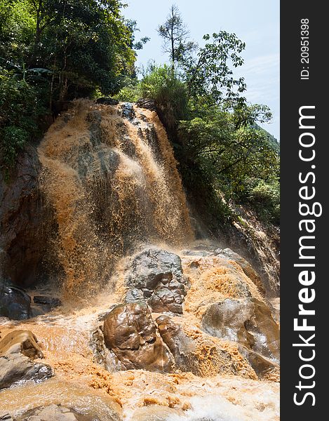 A clayey waterfall with brown water running fast from the hill. One of the stones resembles a human face.