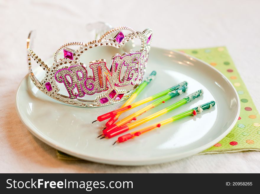 A sparkly rhinestone tiara with silver and purple beads that spells out Princess sits on a blue plate next to used rainbow birthday candles that have been blown out. A sparkly rhinestone tiara with silver and purple beads that spells out Princess sits on a blue plate next to used rainbow birthday candles that have been blown out.