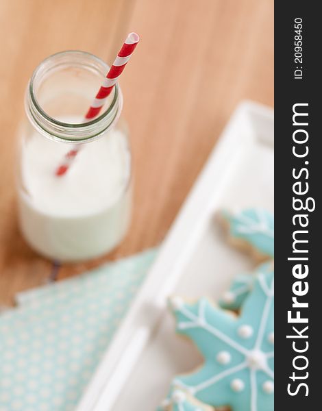 A mini jug of milk with a red and white striped straw sits next to a tray of Christmas blue snowflake bookies. A mini jug of milk with a red and white striped straw sits next to a tray of Christmas blue snowflake bookies.