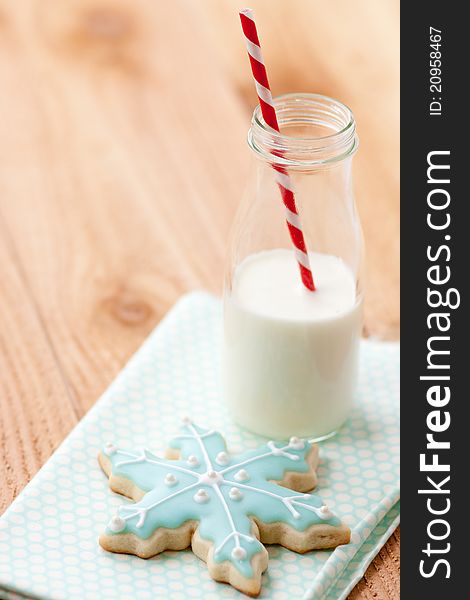 One blue snowflake sugar cookie rests on blue napkin next to a small farm bottle of milk with a red and white striped straw. One blue snowflake sugar cookie rests on blue napkin next to a small farm bottle of milk with a red and white striped straw.