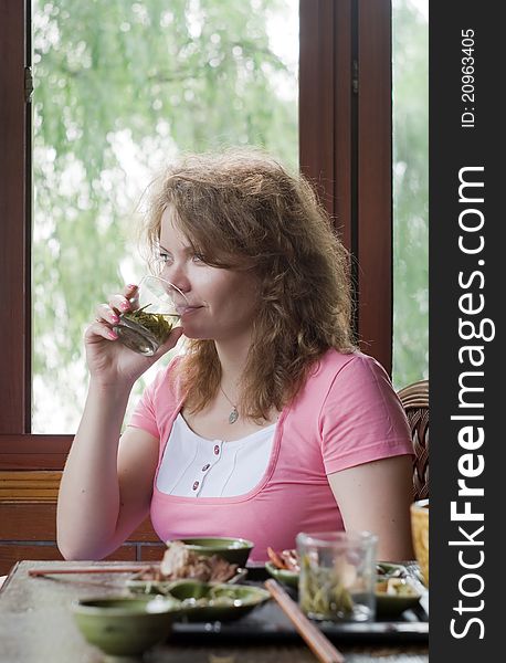 Portrait image of woman which is drinking the green tea opposite the window with beautiful view on the nature. On the foreground is Chinese food on the table. Young woman is smiling. Portrait image of woman which is drinking the green tea opposite the window with beautiful view on the nature. On the foreground is Chinese food on the table. Young woman is smiling.