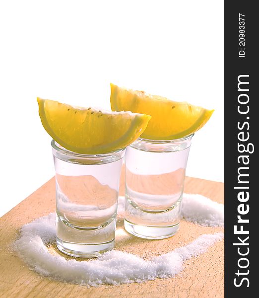 Two glasses filled with tequila, with slices of lemon on the top of it, all surronded with salt, isolated in white. Two glasses filled with tequila, with slices of lemon on the top of it, all surronded with salt, isolated in white