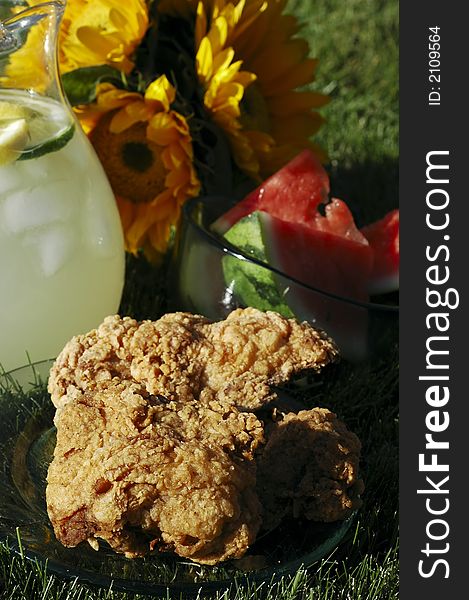 Pitcher of lemonade, plate of chicken, bowl of watermelon, vase of sunflowers on grass in summer afternoon sunlight. Pitcher of lemonade, plate of chicken, bowl of watermelon, vase of sunflowers on grass in summer afternoon sunlight