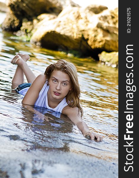 Redheaded girl in a wet white T-shirt sitting in the water and playing with mud