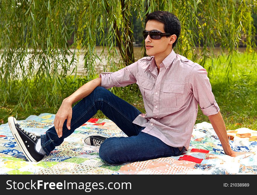 A portrait of a handsome young man, sitting outside on a picnic blanket on a sunny day, with a willow tree behind him. A portrait of a handsome young man, sitting outside on a picnic blanket on a sunny day, with a willow tree behind him