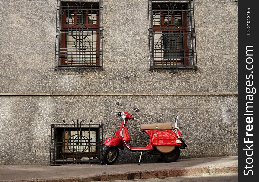 A red moped parked on a sidewalk in front of a building. A red moped parked on a sidewalk in front of a building