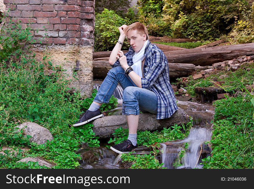 Young human is siting among greenery, old logs and small beautiful waterfall