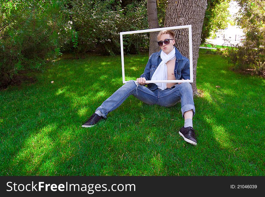 Young human is siting on the green grass with white frame in his hand, he has blue goggles and jean jacket and white scarf he is funny. Young human is siting on the green grass with white frame in his hand, he has blue goggles and jean jacket and white scarf he is funny