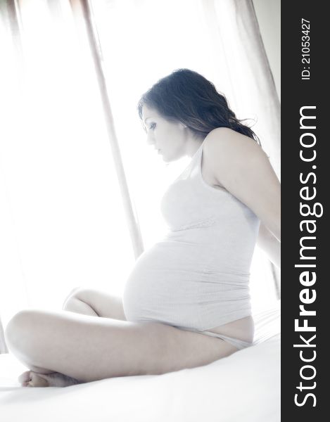 Pregnant woman sitting on the bed with window and curtain in the background with backlighting. Pregnant woman sitting on the bed with window and curtain in the background with backlighting