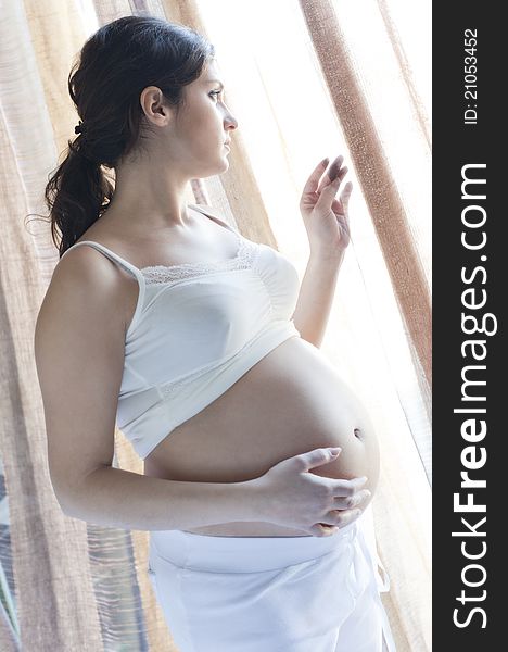 Pregnant woman with white pants and white vest that looks out the window. Pregnant woman with white pants and white vest that looks out the window