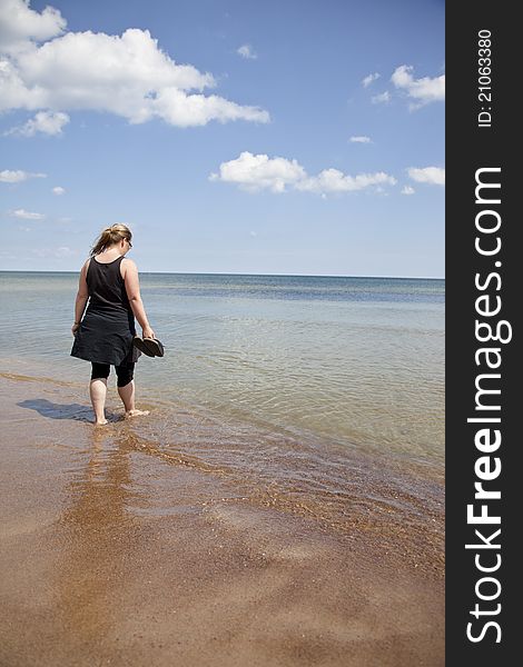 Woman standing at a beach with feet in the water and shoes in her hand. She is looking down into the water. Woman standing at a beach with feet in the water and shoes in her hand. She is looking down into the water.