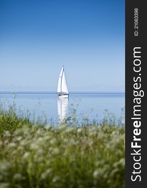 Sailboat on calm water with reflection in water surface. Green grass from land in foreground. Sailboat on calm water with reflection in water surface. Green grass from land in foreground.