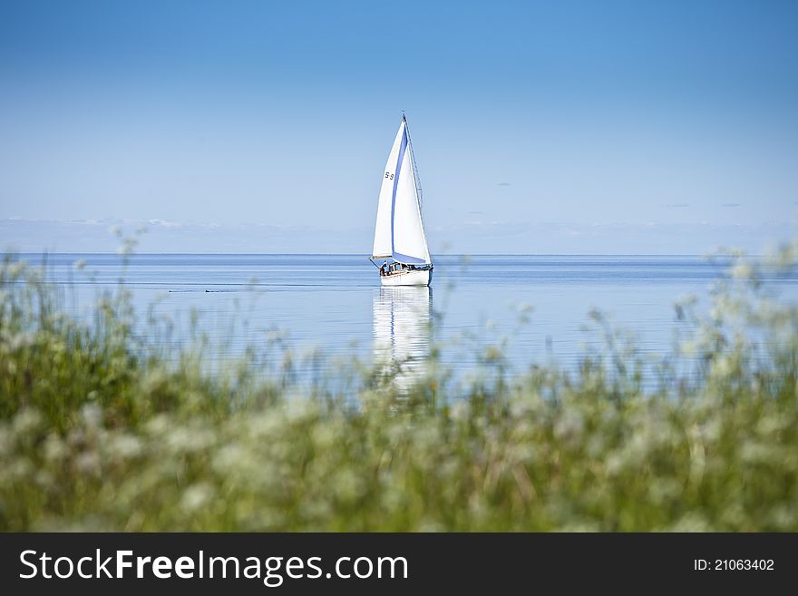 Sailboat on calm water with reflection in water surface. Green grass from land in foreground. Sailboat on calm water with reflection in water surface. Green grass from land in foreground.