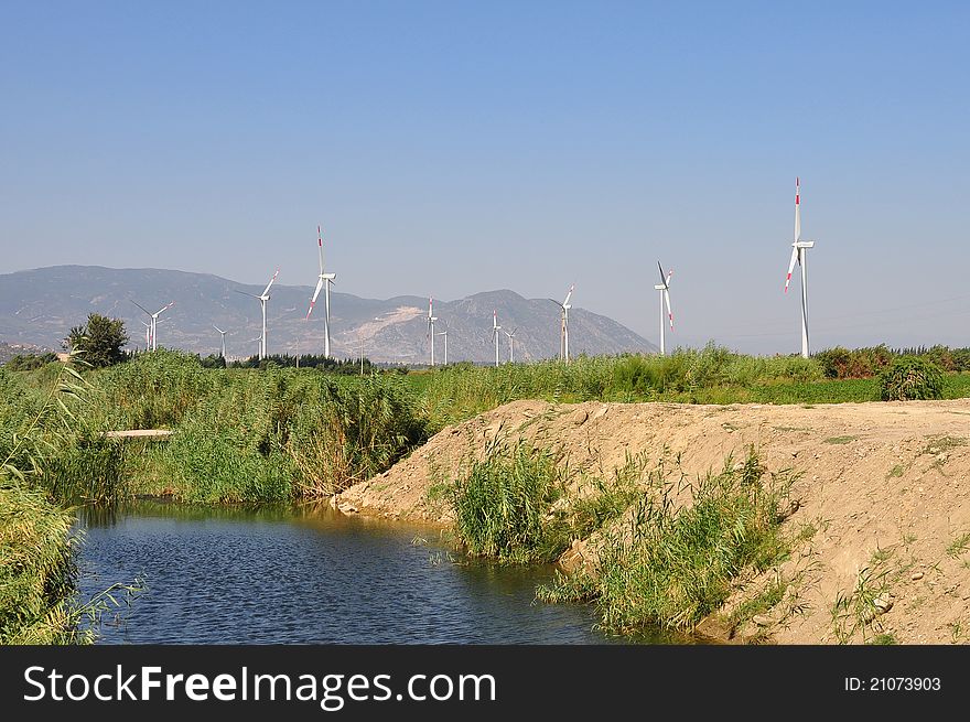 Thirteen white wind turbines in windy valley covered with greenery under bright sun and blue sky, near a creek, with mountains in the background. Soke, Turkey. Thirteen white wind turbines in windy valley covered with greenery under bright sun and blue sky, near a creek, with mountains in the background. Soke, Turkey.