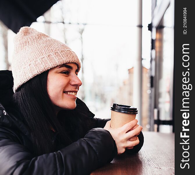 Girl smiling, holds paper cup of coffee in hand near cafe. Blurred background