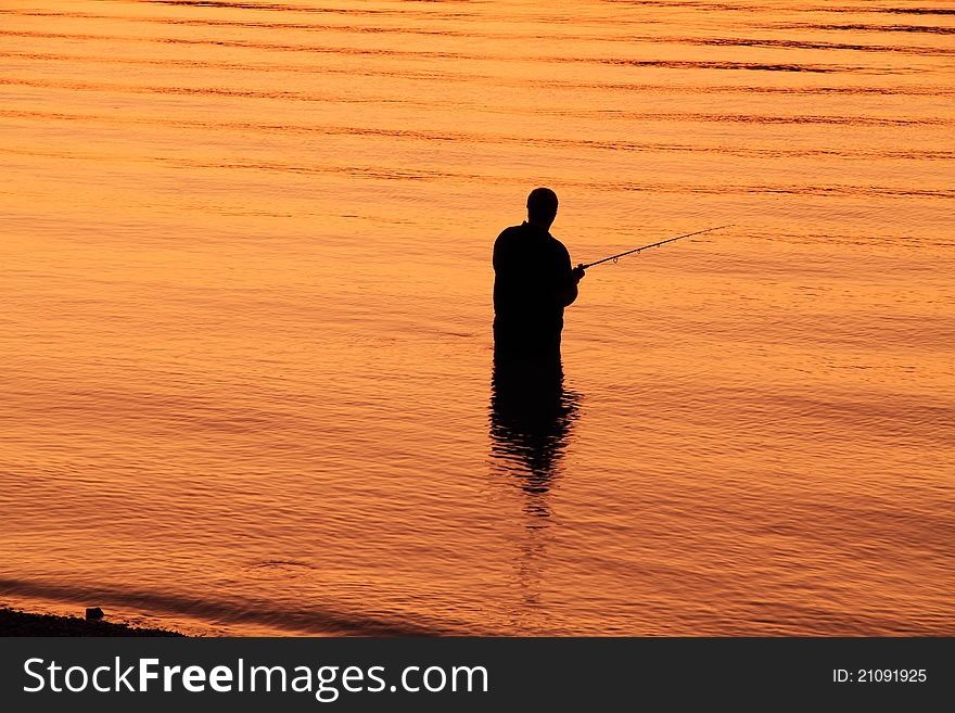 A lone fisherman is silhouetted against the water which is golden orange because of the setting sun. A lone fisherman is silhouetted against the water which is golden orange because of the setting sun.