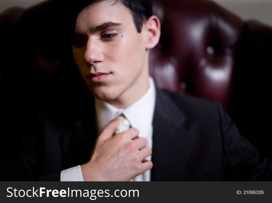 Portrait of a pensive young man dressed in a business suit, touching his tie, seated in a plush leather chair and looking down camera left. Portrait of a pensive young man dressed in a business suit, touching his tie, seated in a plush leather chair and looking down camera left.