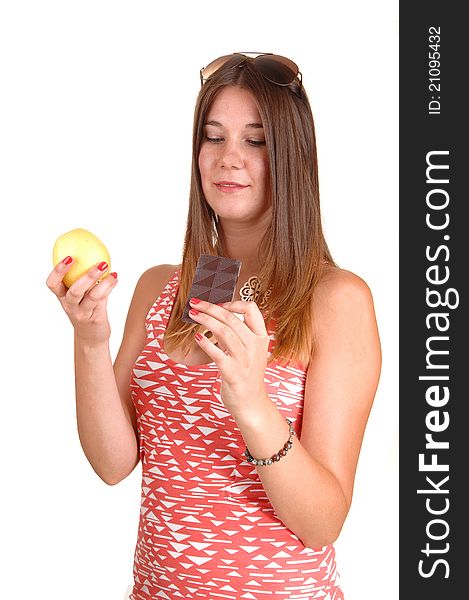 A young woman in a pink dress and sunglasses on her head can not decide to eat an apple or chocolate, in white background. A young woman in a pink dress and sunglasses on her head can not decide to eat an apple or chocolate, in white background.