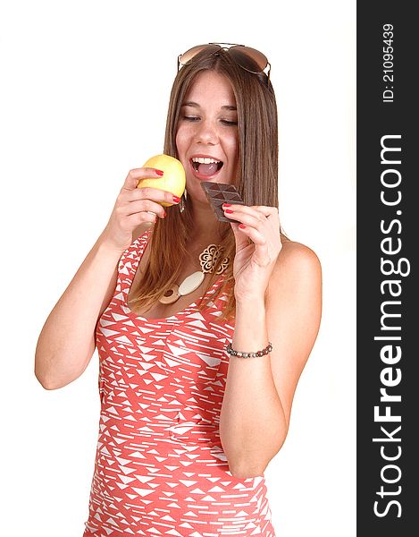 A young woman in a pink dress and sunglasses on her head can not decide to eat an apple or chocolate, in white background. A young woman in a pink dress and sunglasses on her head can not decide to eat an apple or chocolate, in white background.